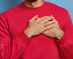 Mann mit rotem Pullover vor blauem Hintergrund, der die Hände vor sein Herz hält